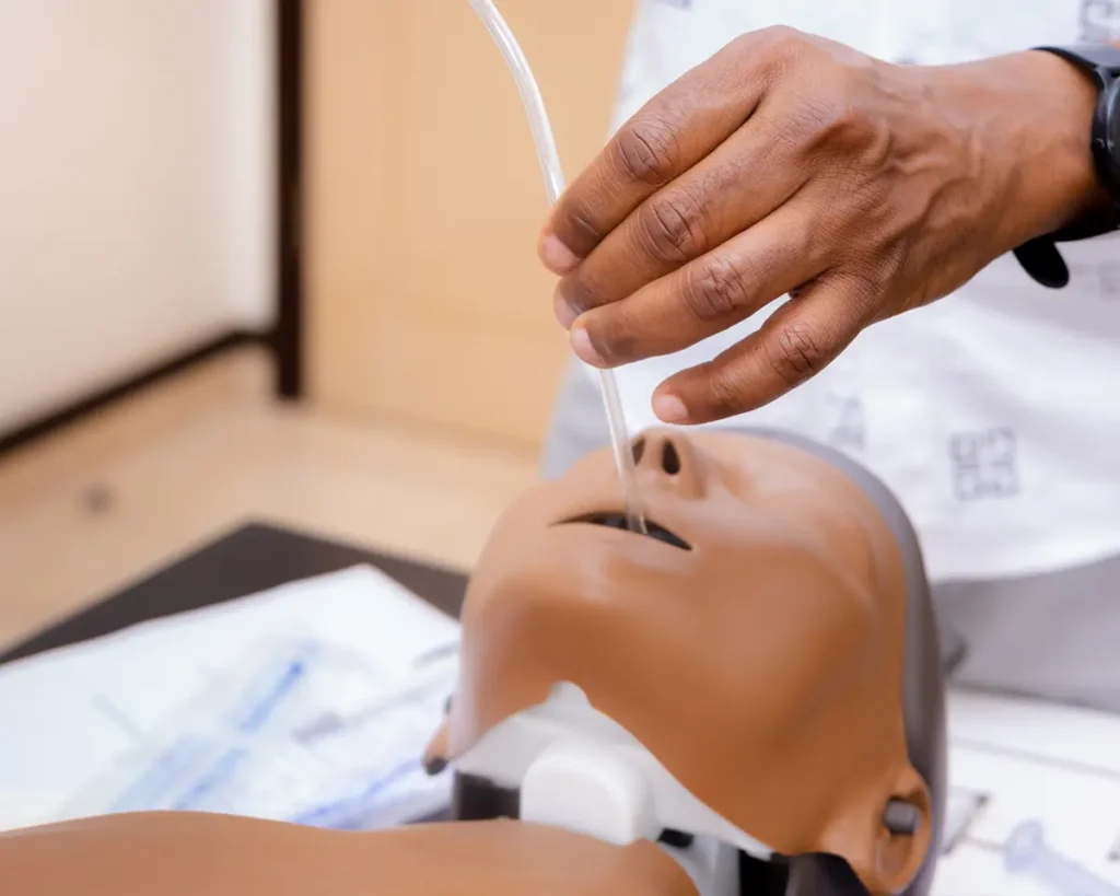 Close-up image showing a person using the Little Anne manikin, with a tube visibly inserted in the manikin's mouth.