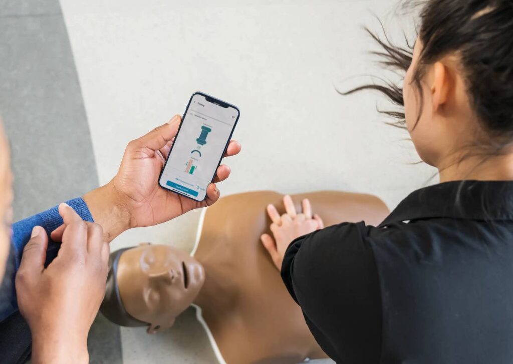 A person is training CPR on a manikin, while another person holds a mobile phone which is visibly showing the QCPR app analysing their performance.